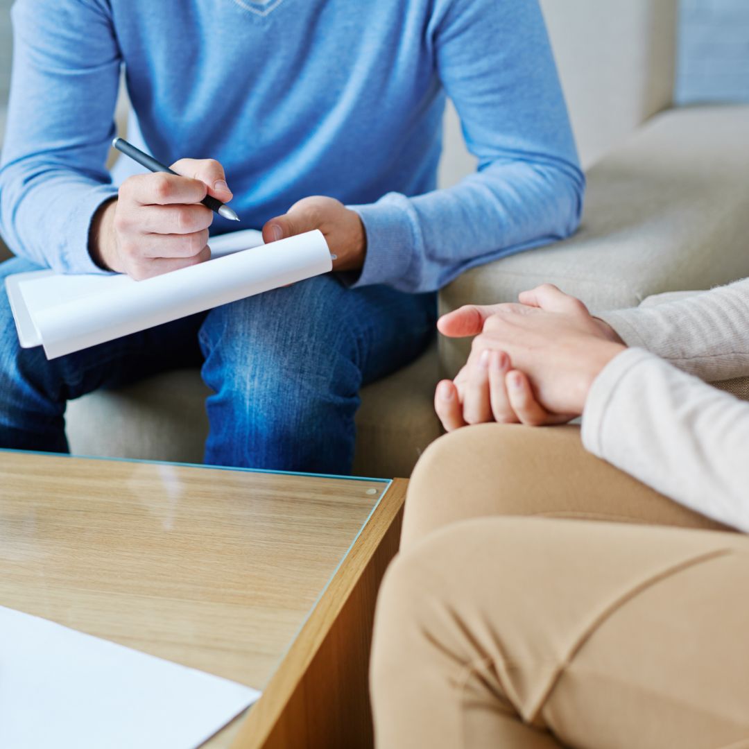 Close up of two individuals sitting closely on two chairs. One individual is facing the camera and is wearing a blue shirt, holding a pad of paper and a pen in their other hand. The person in the blue shirt is facing the other individual. The other individual is wearing a white shirt and brown pants. The person in the white shirt has their hands together in their lap.